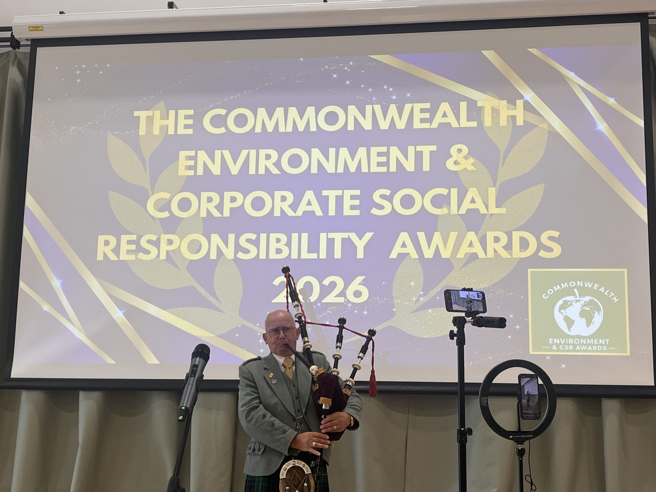 Bagpipe player stands infront of a screen which says: 'The Commonwealth Environment & Corporate Social Responsibility Awards 2026'